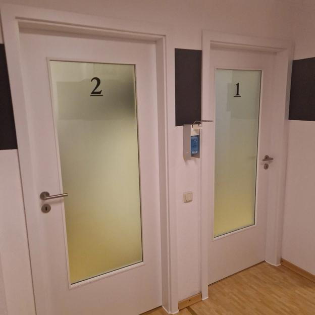 Zwei weiße Praxistüren mit Glaseinsatz und schwarzer Nummerierung für Räume.