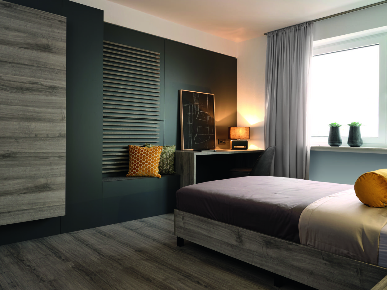 Moderne Schlafzimmeransicht mit einem Bett, Schreibtisch, Sessel, Regalen und Pflanzen vor einem Fenster.